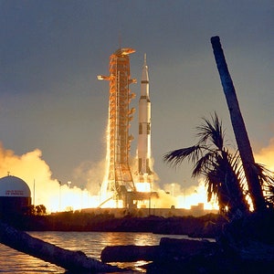 May include: A white and orange rocket launches from a launchpad at night. The rocket is surrounded by smoke and fire. The launchpad is located near a body of water.