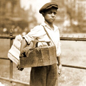 May include: A young boy wearing a cap and a white shirt carries a wooden box with a shoe repair kit. The box has a strap and is slung over his shoulder. He is standing in front of a metal fence.