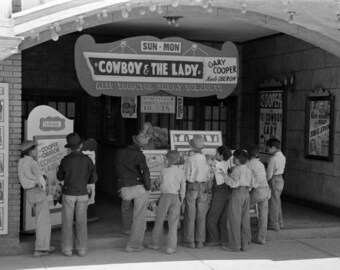 May include: A black and white photo of a movie theater marquee advertising the film "Cowboy & The Lady" starring Gary Cooper and  Madeline Carroll. The marquee also advertises the admission price of 10 or 25 cents. A group of people are standing in front of the theater, some looking at the marquee and others looking at the ticket booth.