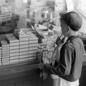 Könnte beinhalten: Ein junger Mensch mit einer Baseballkappe betrachtet eine Vitrine mit Spielzeug-Kapselpistolen und Schachteln mit Kapseln. Die Vitrine befindet sich hinter einer Glasscheibe. Die Schachteln mit Kapseln sind hoch gestapelt und mit "Cap Pistols" beschriftet.