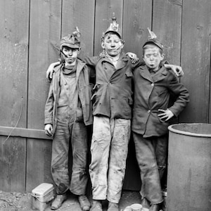 May include: Black and white photograph of three young boys, likely coal breakers, posing together. They wear work clothes, caps with lights, and have coal dust on their faces. The image is a historical depiction of child labor.