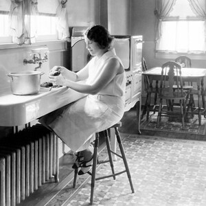 May include: A woman in a white dress sits on a stool in a kitchen, preparing food at a sink. She is holding a potato in her hand and there is a pot on the counter. The kitchen has a tiled floor, a radiator, and a window with curtains.