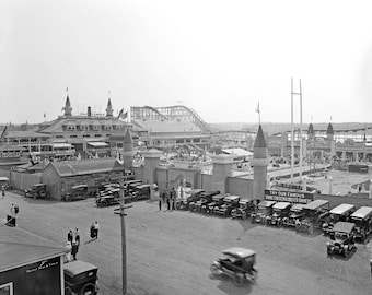 May include: A black and white photo of a crowded amusement park. The photo shows a large building with a roller coaster in the background. There are many people walking around and cars parked in the lot. A sign reads "Try Our Famous Shore Lobster & Oyster Dinners".