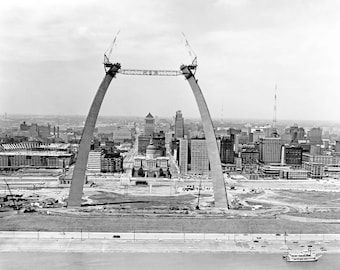 Puede incluir: Una fotografía aérea en blanco y negro del Arco Gateway en St. Louis, Missouri, en construcción. El arco está hecho de acero y está siendo ensamblado por grúas. El horizonte de la ciudad es visible en el fondo.