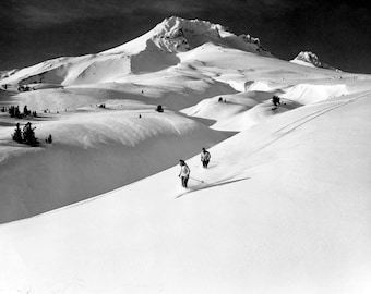 Könnte beinhalten: Zwei Skifahrer in schwarz-weißer Kleidung fahren einen schneebedeckten Berghang hinunter, im Hintergrund ein Berggipfel.