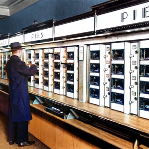 May include: A vintage vending machine with multiple rows of glass doors, each containing a different type of pie. The machine is labeled "PIES" in black lettering. A person in a dark coat is standing in front of the machine, looking at the selection.