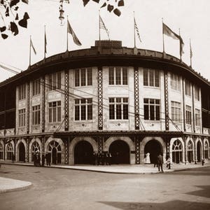 May include: A black and white photo of a large, brick building with arched entrances. The building has a curved roof and many windows. There are several people walking in front of the building.