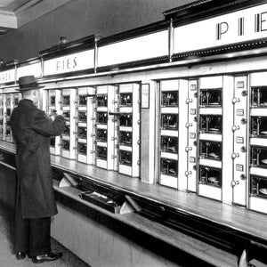 May include: A black and white photo of a vintage vending machine with a selection of pies and cakes. The machine has multiple rows of glass doors with food items visible inside. A person is standing in front of the machine, looking at the selection.