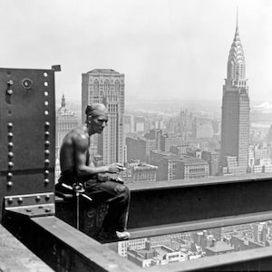 May include: A black and white photo of a construction worker sitting on a beam high above the city. The worker is wearing a white undershirt and blue jeans. The Chrysler Building is visible in the background.