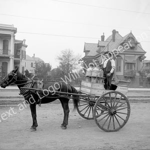 May include: A black and white photo of a horse-drawn cart with a man standing on the back. The cart is carrying several large milk jugs. The cart is on a dirt road in front of a house. The text on the cart reads 