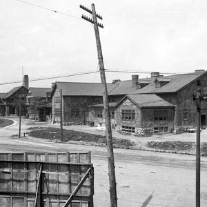 May include: A black and white photo of a large, wooden building with a gabled roof. The building appears to be under construction, with some of the windows boarded up. There are power lines running across the front of the building.