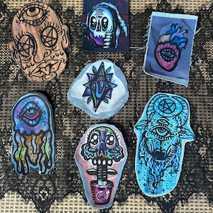 May include: A collection of seven colorful, hand-drawn patches with various occult and surrealist designs. The patches feature images of skulls, eyes, hearts, and other mystical symbols, with a variety of colors and styles.