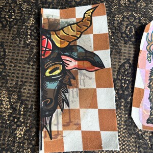 May include: A rectangular patch featuring a stylized illustration of a goat's head with large horns, a red eye, and a checkered background in brown and white. The artwork is detailed with black outlines and various colors.