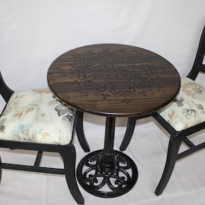 May include: A black and brown round table with a decorative design on the top. Two black chairs with floral patterned cushions are placed around the table.