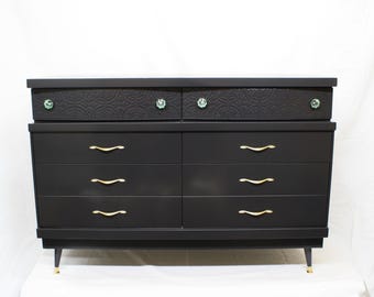 May include: A black dresser with six drawers and gold handles. The top two drawers have a textured surface and green glass knobs.