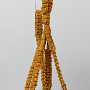 May include: A mustard yellow macrame plant hanger featuring intricate knotted cord patterns, a central suspension loop, and multiple braided support strands that extend downward to cradle a plant container with dark green leaves.