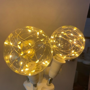 May include: A decorative lamp featuring two clear glass globes filled with warm white LED string lights. The globes are mounted on white fixtures with silver accents, creating a soft, ambient glow. The lamp is designed for indoor use.