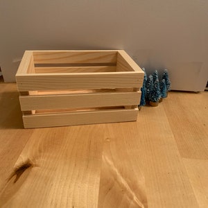 以下が含まれることがあります： 水平スラットを備えた、小さな未完成の木製クレート。 クレートは正方形で、明るい色の木製の表面に置かれています。 小さな青いボトルブラシの木がクレートの右側にあります。