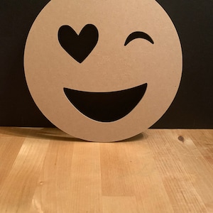 May include: A wooden cutout of a smiling emoji with one eye closed and a heart in the other eye. The emoji is brown and has a white background.