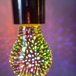 May include: A glass light bulb with a multi-colored 3D firework effect. The bulb features a dark metallic base and emits a vibrant display of yellow, green, and pink light points that radiate outward, creating a colorful pattern on the surrounding area.