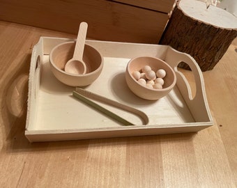 May include: A light-coloured wooden serving tray containing two small wooden bowls, a wooden spoon, a pair of light green wooden tongs, and several small wooden spheres. The tray features cutout handles on the sides and a smooth, natural finish.