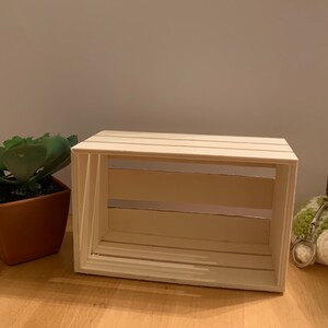 May include: A white wooden crate with a rustic look. The crate has four sides and a bottom, and it is open on the top. The crate is made of wood slats that are joined together with nails. The crate is empty.