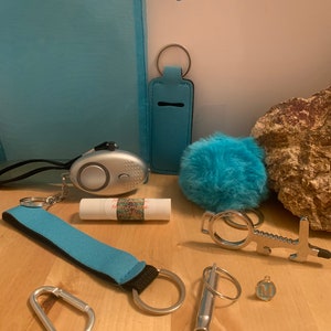 Puede incluir: Una bolsa de malla azul llena de varios artículos para llavero, que incluyen una alarma personal, un silbato, un llavero, un llavero peludo azul, un bálsamo labial y un llavero con una cubierta de tela azul.