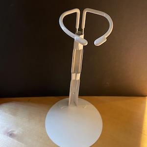 May include: A white metal doll stand with a circular base and a curved top. The stand is designed to hold a doll upright. The stand is made of metal and has a simple, functional design.