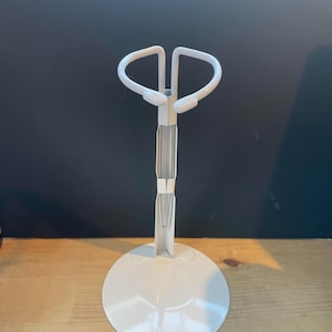 May include: White plastic doll stand with a round base and a curved top for holding a doll.