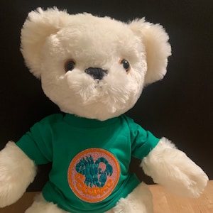 Op de afbeelding: Witte pluche teddybeer die een groen T-shirt draagt met een geborduurde schorpioen en het woord "Scorpio" erop.