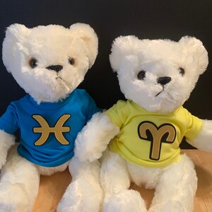 Op de afbeelding: Twee witte pluche teddyberen die blauwe en gele T-shirts dragen met sterrenbeeld symbolen. Het blauwe shirt heeft een gouden Vissen symbool en het gele shirt heeft een gouden Ram symbool.