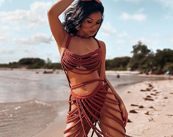 May include: A woman wearing a rust-colored crocheted bikini top and skirt on a sandy beach. The top and skirt are made of thick rope and have a fringe detail.