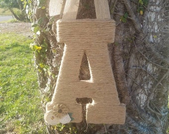 May include: A large letter A made of twine with a burlap bow and white flowers attached. The letter is hanging from a tree.
