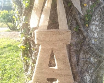 May include: A large letter A made of brown twine with a burlap bow and a small flower embellishment. The letter is hanging from a tree.