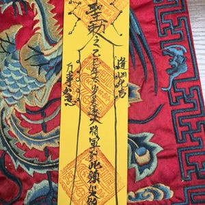 Puede incluir: Un papel amarillo con caligrafía negra y sellos rojos, sobre una tela bordada roja con un diseño de fénix azul y verde. El papel tiene líneas verticales y texto.