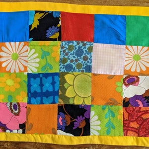 May include: A patchwork table runner with a yellow border. The squares display floral and geometric patterns in blue, green, orange, and purple. The runner is likely made of fabric.