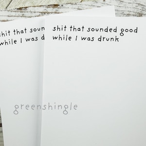 May include: Two white greeting cards with black handwritten text that reads "shit that sounded good while I was drunk" and "shit that sounded good while I was drunk". The cards are on a white wooden surface.