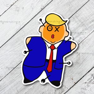 May include: A cartoon sticker of a figure in a blue suit and red tie, with yellow hair and an orange face. The figure has pins stuck in it, with one eye closed and an X over the other. The sticker is on a white wooden surface.