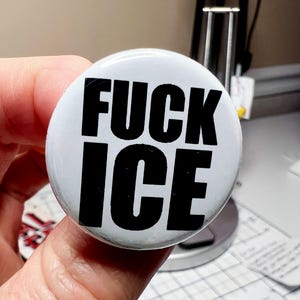 May include: A white button with the words "FUCK ICE" in bold, black capital letters. The button is round and held in a hand.