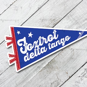 May include: A pennant-shaped sticker with a blue background and white text that reads "Foxtrot delta tango." The pennant has a red border with white stitching and white stars. The sticker is on a white wooden surface.