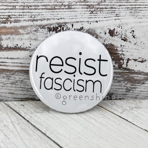 May include: White round button with black text that reads "resist fascism" and a small copyright symbol with the text "greenshin" below.