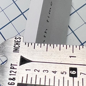 Pu&ograve; includere: Un primo piano di un righello di metallo con segni in pollici. Il righello &egrave; contrassegnato con numeri da 1 a 7. Il testo "6 & 12 PT. INCHES" &egrave; stampato sul righello.