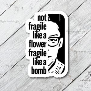 May include: Black and white sticker with a quote that reads "not fragile like a flower fragile like a bomb". The quote is next to a black and white illustration of a person wearing glasses.