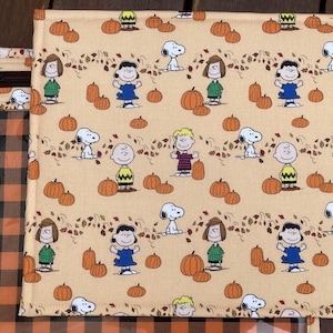 May include: A brown and black checked fabric with a beige fabric panel featuring a repeating pattern of Peanuts characters and pumpkins. The Peanuts characters include Charlie Brown, Snoopy, Lucy, Linus, and Sally. The fabric is ideal for crafting projects such as tote bags, cushions, and quilts.