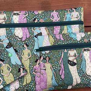 May include: Three green zippered pouches with a floral pattern featuring women in dresses and a green background. The pouches are different sizes and have a zipper closure.