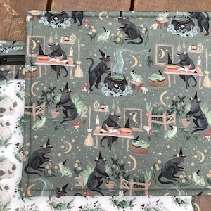 May include: A green fabric with a repeating pattern of black cats, cauldrons, and stars. The cats are wearing witch hats and holding wands. The fabric is ideal for crafting projects such as tote bags, cushions, and quilts.