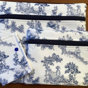 Op de afbeelding: Een set stoffen zakjes met toile-de-jouy-patroon in verschillende maten. De witte stof heeft een blauwe pastorale scène. Elk zakje heeft een zwarte ritssluiting. Een klein zakje heeft een blauwe knoopsluiting.