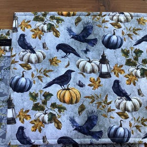 May include: A rectangular fabric item featuring a repeating pattern of black crows, pumpkins, butterflies, and autumn leaves on a light blue background. The pumpkins are white, orange, and blue. A decorative lantern is also featured.