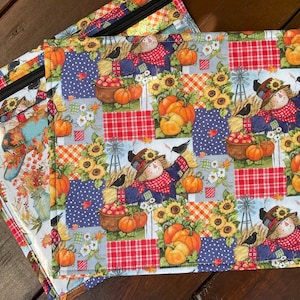 Peut inclure: Deux pochettes rectangulaires en tissu avec des fermetures éclair noires. Les pochettes présentent un motif patchwork avec des épouvantails, des citrouilles, des tournesols et des moulins à vent sur le thème de la récolte d'automne. Le fond est une surface en bois.