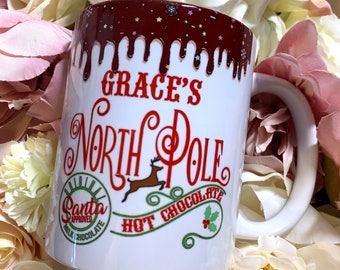 May include: White ceramic mug with a red and white design. The mug has a red dripping chocolate design with white sprinkles and the text "Grace's North Pole Hot Chocolate" in red. The mug also has a red and white circle with the text "Original Santa Approved Milk Chocolate" in white.